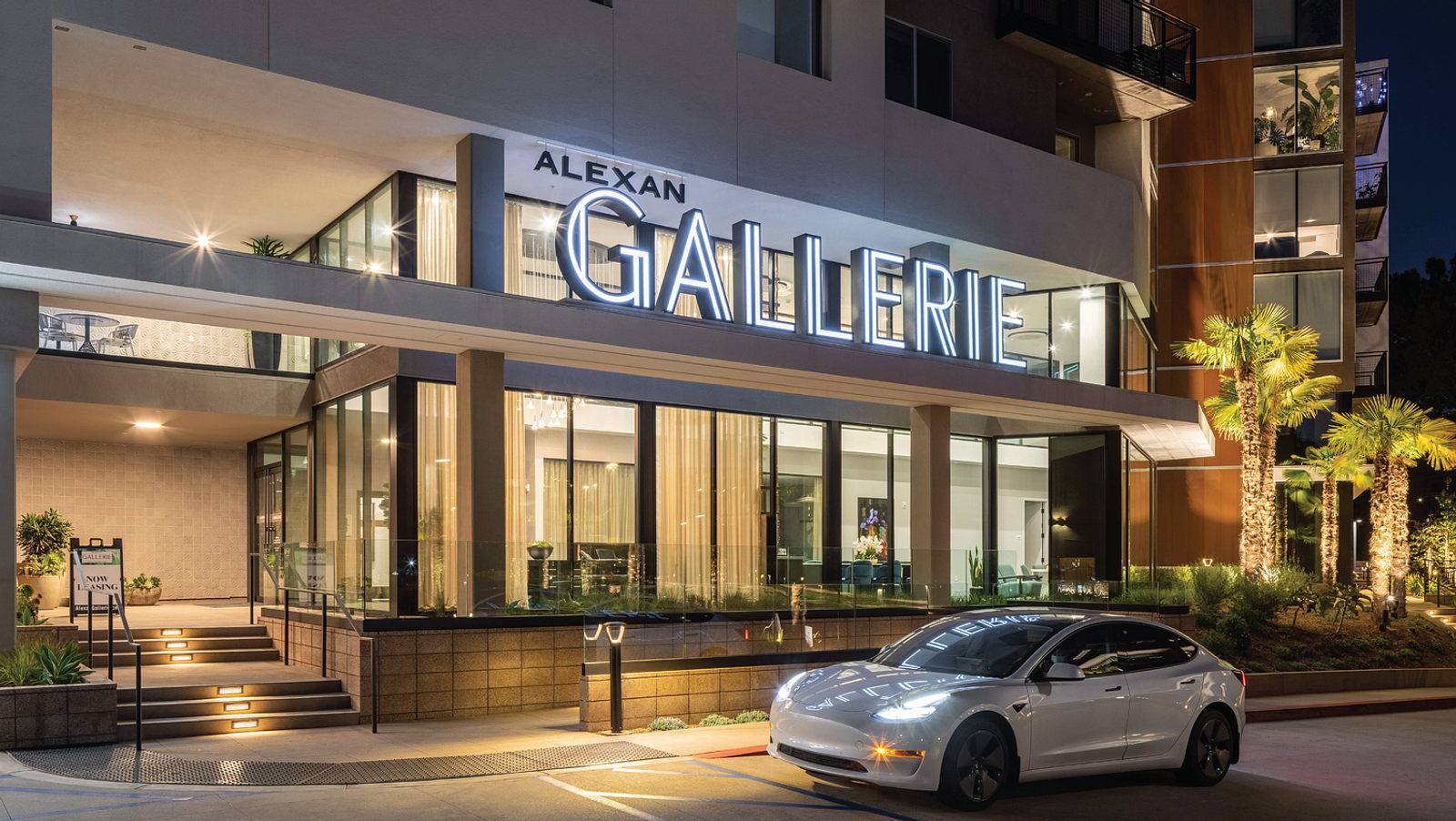 Building ID sign with the letters of Gallerie lit up with white light on the exterior of a building with a white tesla parked in front.