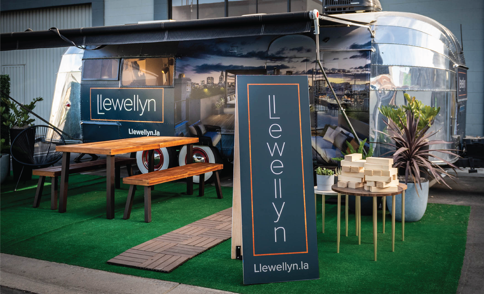 A free standing blue sign with the Llewellyn logo place on top of green turf with a picnic table slightly behind the sign and a food trailer in the background