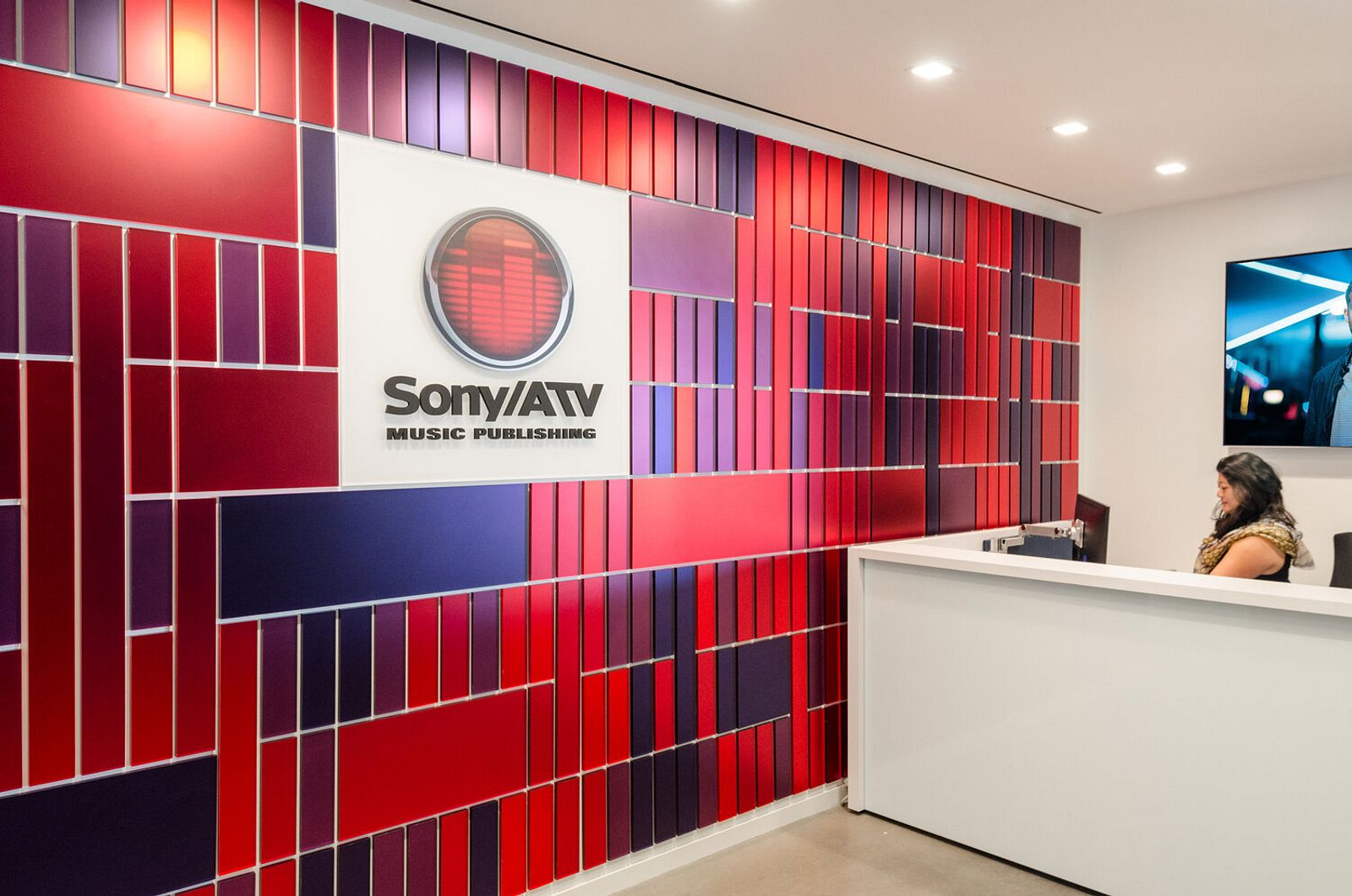 An interior wall made up of blue and different shades of red panels of various sizes with a logo in the center and a women sitting behind a white receptionist desk to the right.