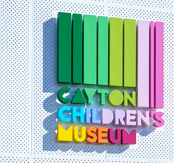 Colorful building id signage of Cayton's logo mounted on a white wall with small holes for texture with a shadow being cast from the sign.