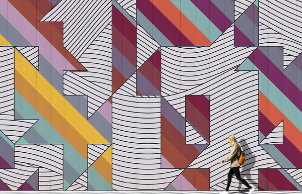 Person walking by a huge stripped colorful mural with large shapes of black and white stripes cut out of it.