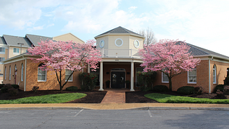 V-shaped brick building with brick sidewalk leading to entrance and pink flowering trees on either side of the entrance walkway