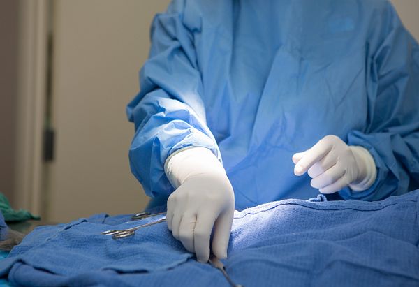 Surgeon with blue gown and white gloves picking up a spay hook from a blue drape.