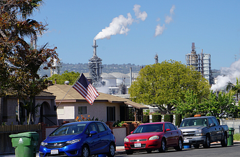 Cars in front of houses in residential neighborhood, with large refinery in near background