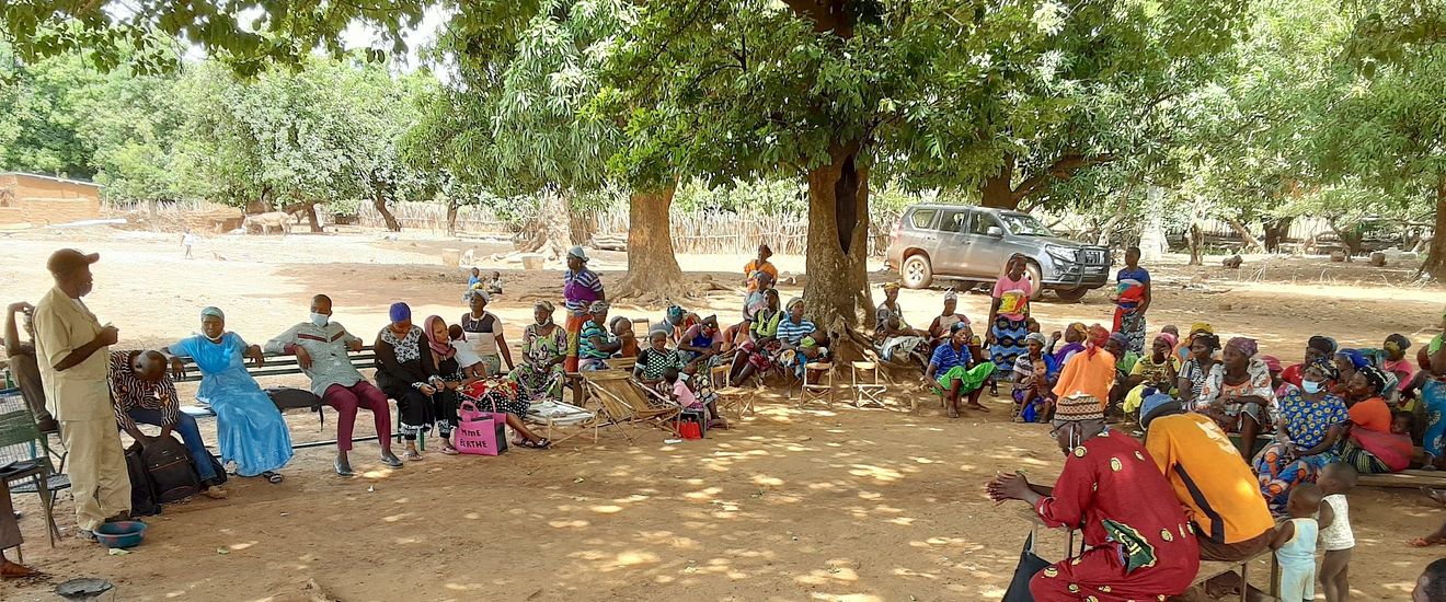 A man stands on the left speaking to a group of people sitting in a semi-circle outside under trees.