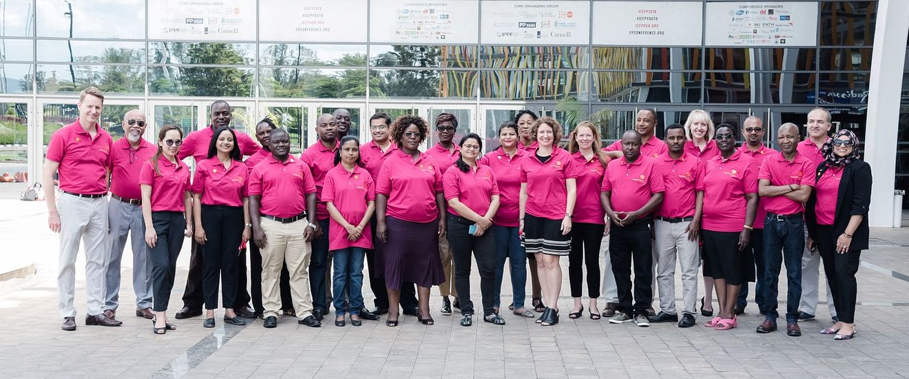 A group fo 27 people, all wearing magenta polo shirts, stand together outside of a conference center.