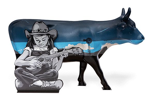 Right facing cow spilt into 3 blue + black colors, top navy, middle is with a outdoor scene with white clouds, mountains and a windmill. Bottom is painted black. A young girl sitting cross-legged in front wearing overalls and cowboy hat looking down/playing a ukulele.