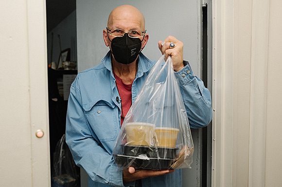 A photo of client, Carlo, wearing a black mask and holding meals from God's Love at his front door.