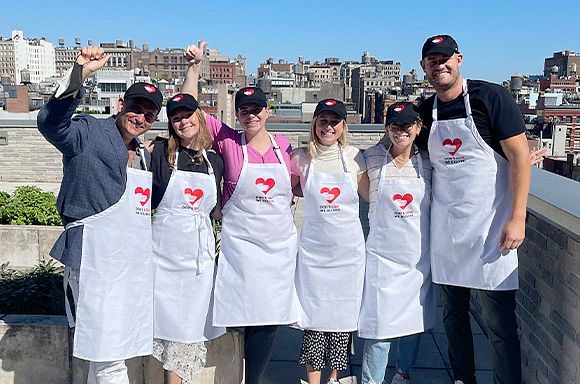 A group photo of volunteers from the Points Guy on the God's Love rooftop. The group of six are smiling and waving at the camera on a sunny day while wearing God's Love hats and aprons.