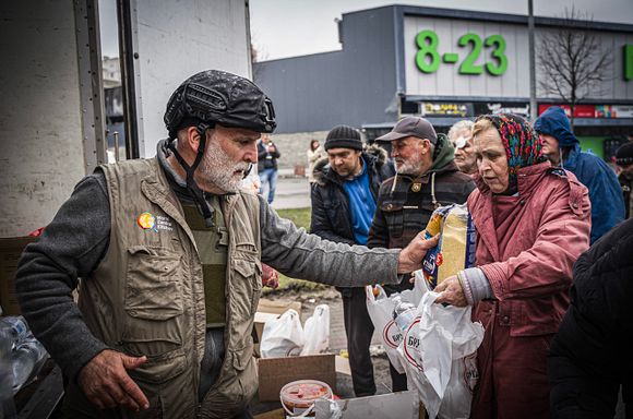 A photo of Chef Jose Andres in a vest and helmet handing out food to people in Ukraine.