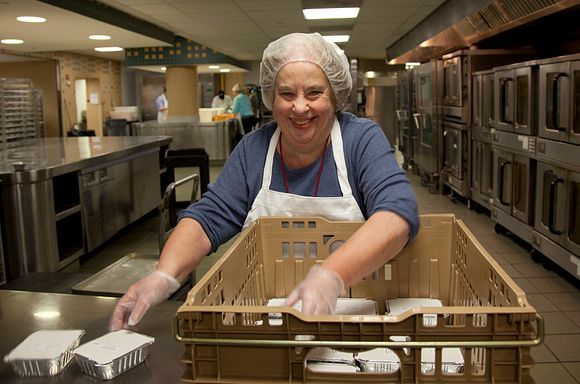 Volunteer Deborah Corenthal wearing a hairnet with a brown crate filled with dessert trays in front of her