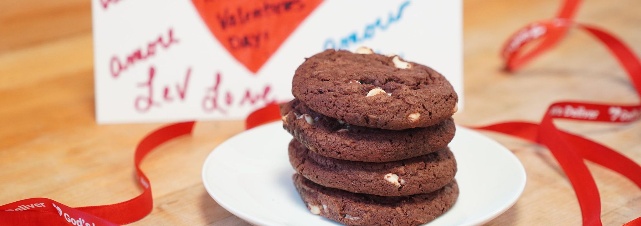 A photo of red velvet cookies stacked on a white plate. In the background there is a Valentine's Day card.