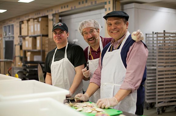 A photo of three volunteers in the God's Love kitchen smiling together and looking at the camera. The volunteers are wearing hairnets and aprons and are peeling potatoes.
