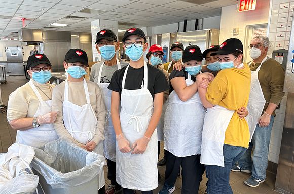 A group photo of students and chaperones from Stephen Gaynor School in the God's Love kitchen posing together in aprons and hats.