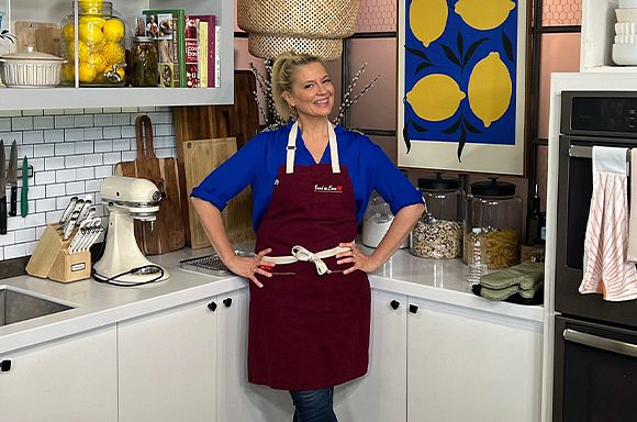 A photo of Chef Amanda Freitag in a kitchen smiling while wearing a blue shirt and a burgundy apron made by Cooks Who Feed.