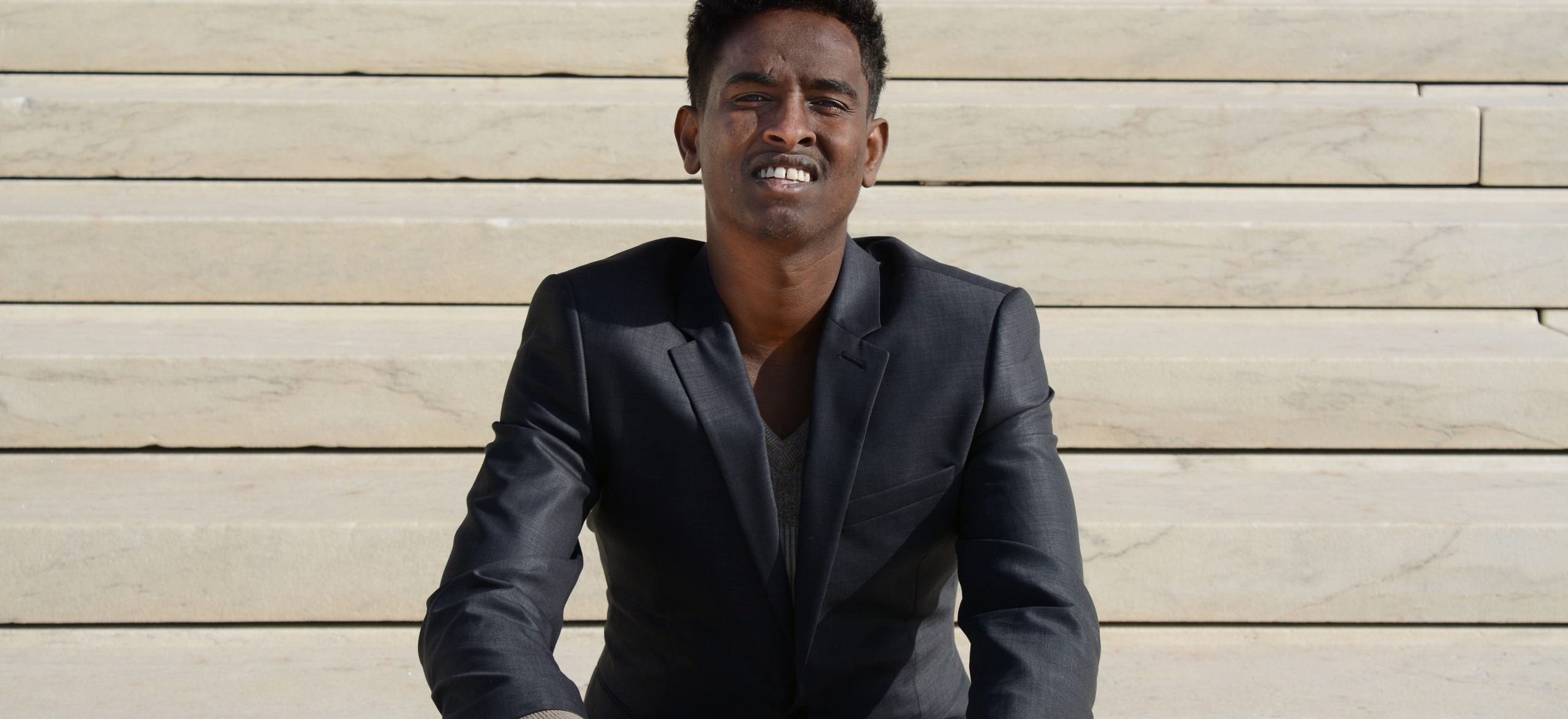 A young Somali man in a black suit jacket sits on the stairs of the U.S. Capitol building.