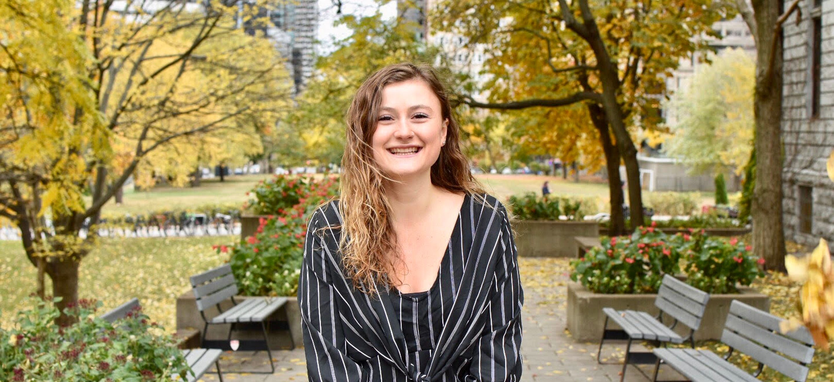 Naomi Santesteban, a student at a Candian IRAP Law School Chapter, smiles into the camera. She has wavy blondish brown hair and wears a black and white striped outfit.In the background, trees with yellow leaves, wooden benches, and stone buildings are visible.