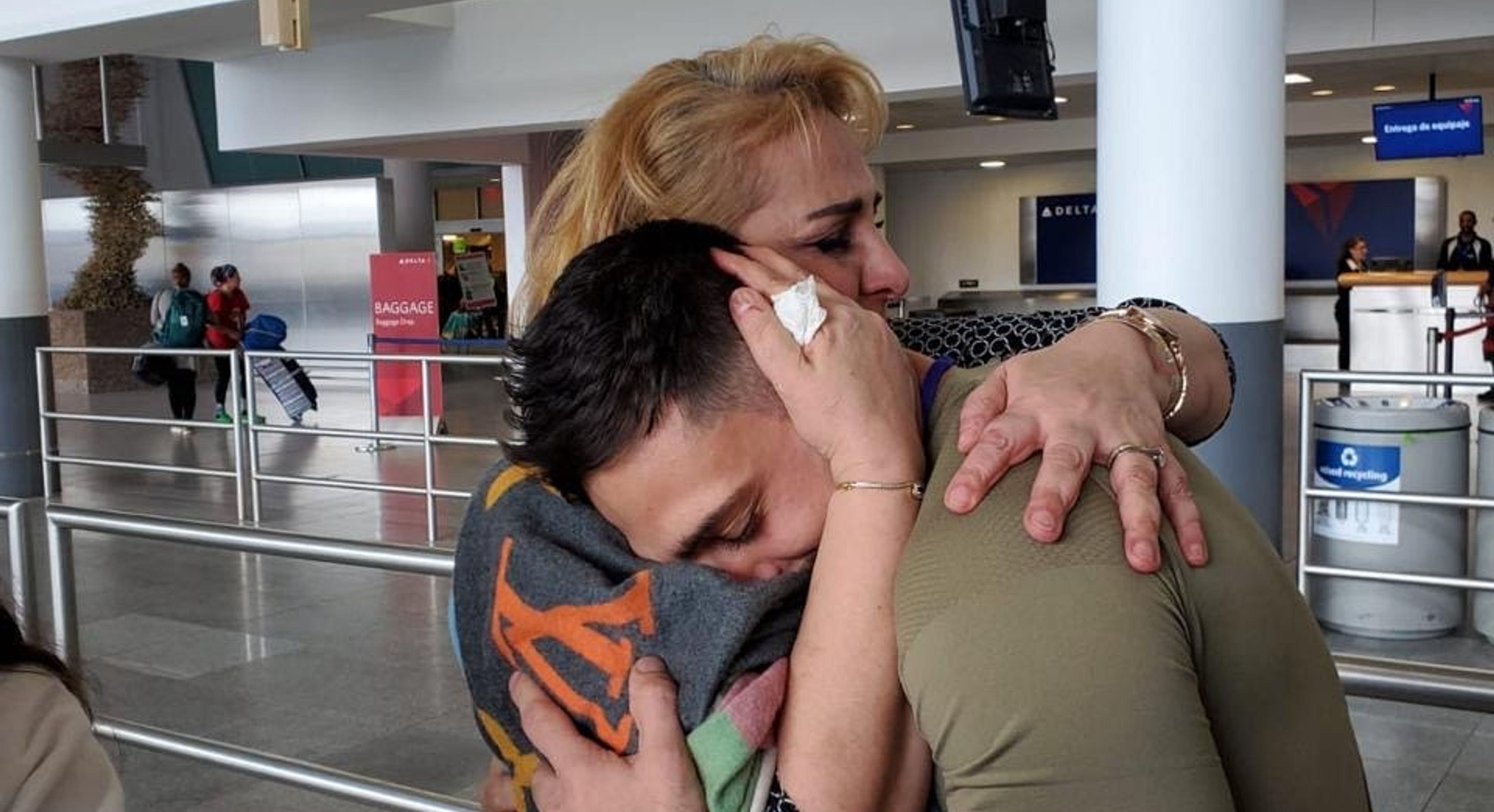 A Syrian mother embraces her son in an airport terminal. The mother has blonde hair and has tears in her eyes. The son nuzzles his face into her shoulder.