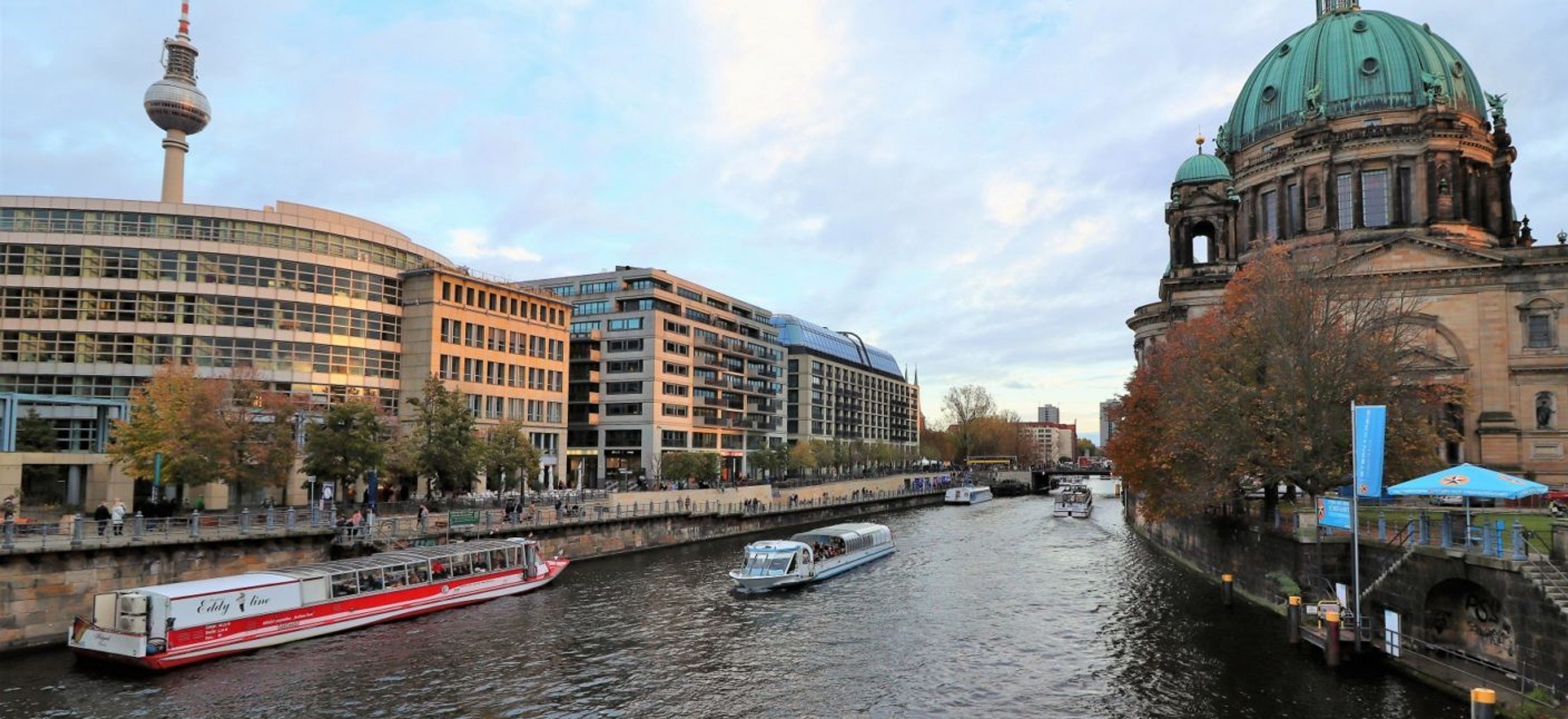 From a bridge over the Spree river in Berlin, Germany, we can see multiple ferry boats on the water. On the left side of the river are modern buildings and the Berlin Television Tower. On the righthand side of the river, is a historic stone building with a green patina dome and a gold spire.
