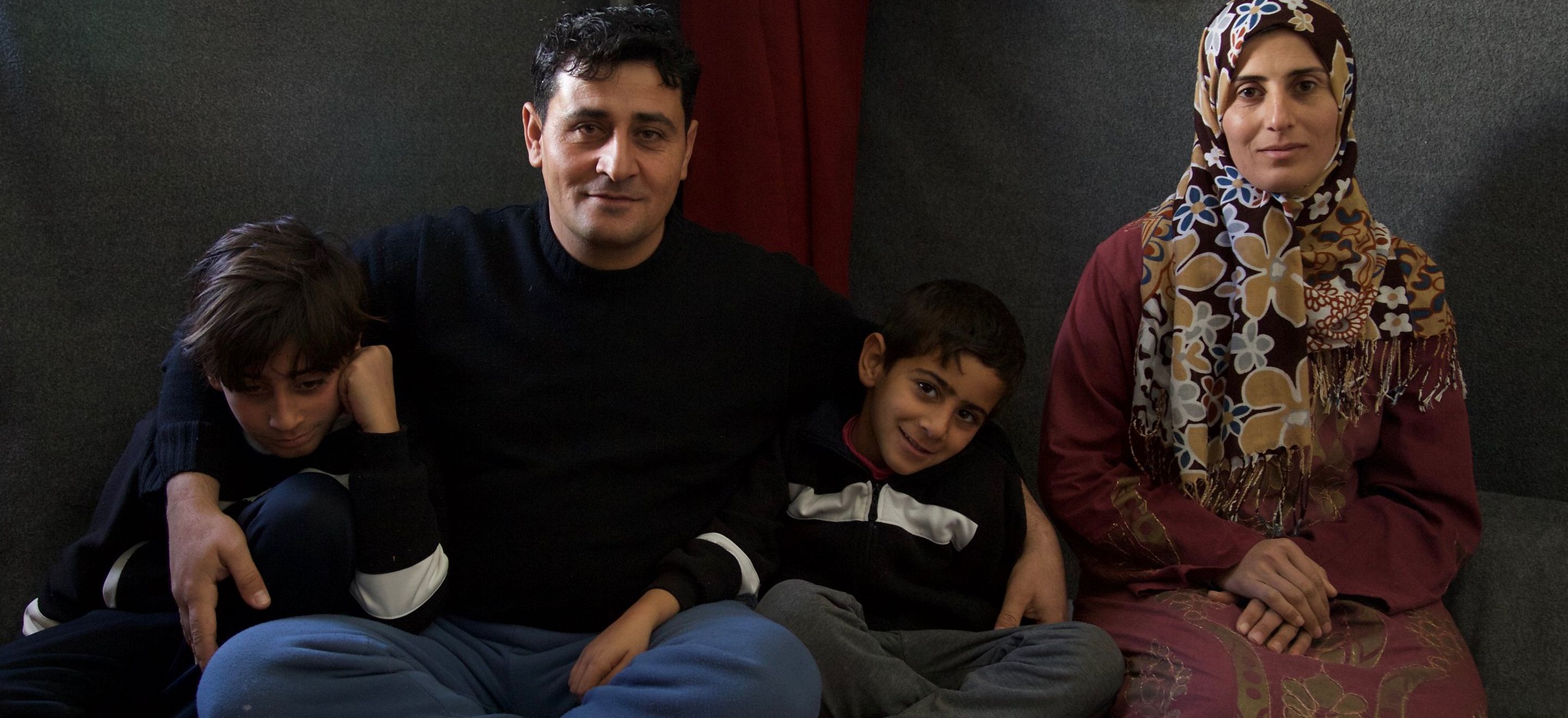 A family of four Syrian refugees - two young sons, a father, and a mother - sit on cushions on the floor.