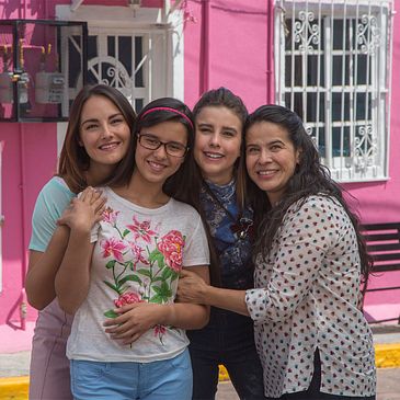 Four main characters of Vencer el Miedo, women of different ages, huddle together posing for a photo. A bright pink building in the background