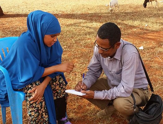 A man writes on a notepad while a women in a blue hijab talks to him