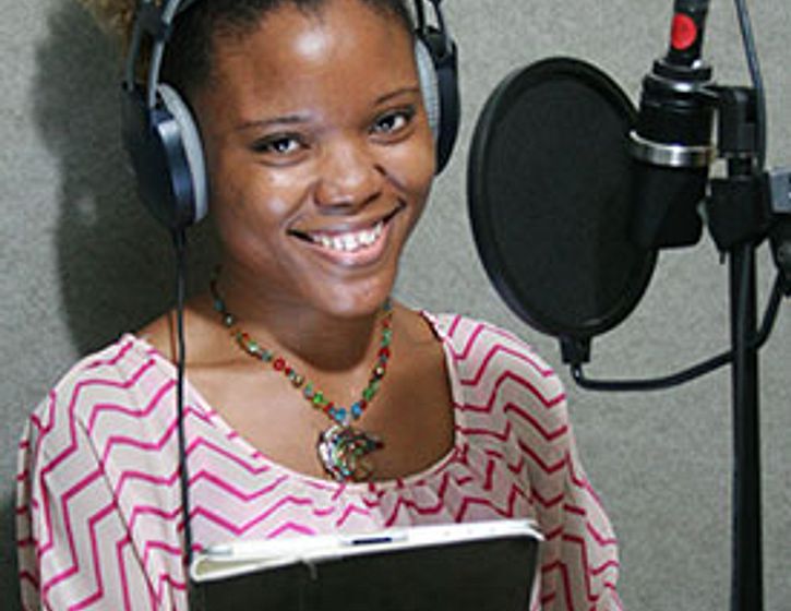 Woman in a recording studio standing in front of a mic, holding a script and wearing headphones