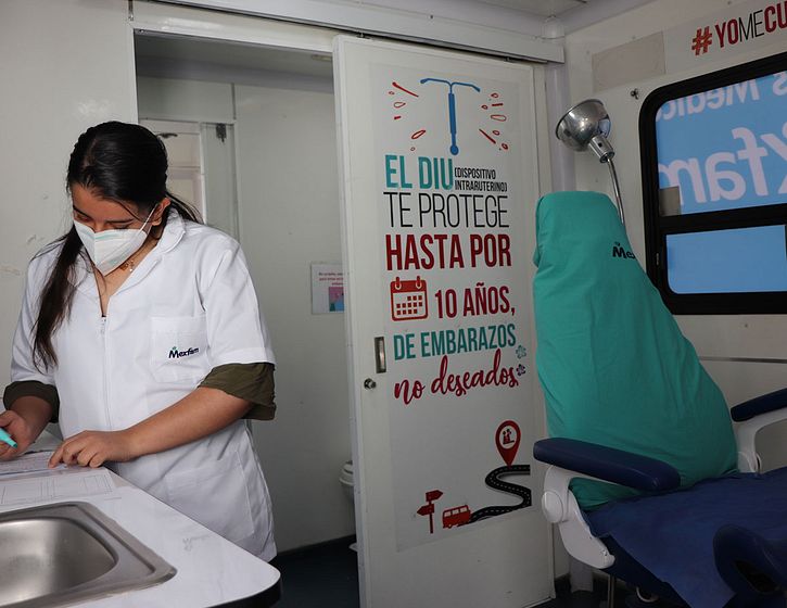 Female OBGYN stands in a mobile health clinic taking notes on a counter. Beside her is a patient chair, sink, and wall art promoting IUDs
