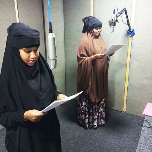 Two women standing in a recording studio. Mics hang down from the ceiling or off the wall. Both women hold scripts and are acting out a scene.