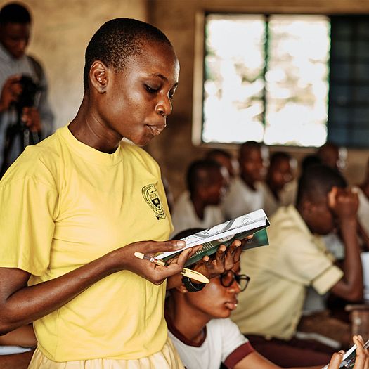 Young woman stands in a school room reading aloud from a book.