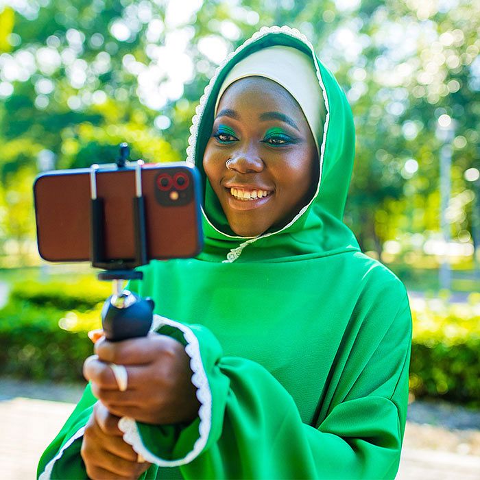 Young woman holds a selfie stick with a phone. She's watching or creating content in an outdoor setting with trees and bushes in the background.