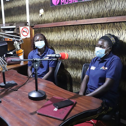 Two PMC-Uganda staff sit in a recording studio desk with microphones pointing toward them.