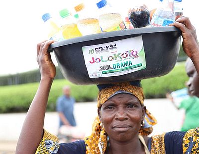 woman carrying a bucket on her head filled with bottled soda drinks. The label on the bucket reads, "Jolokoto."