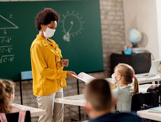 A masked teacher hands a paper to a student