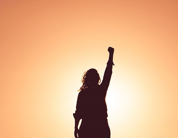 Silhouette of woman standing with her fist in the air. She's blocking the light of the sun but orange sky surrounds her