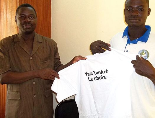 A man presenting a t-shirt prize to another man. The shirt reads, "Yam Yankre, The Choice."