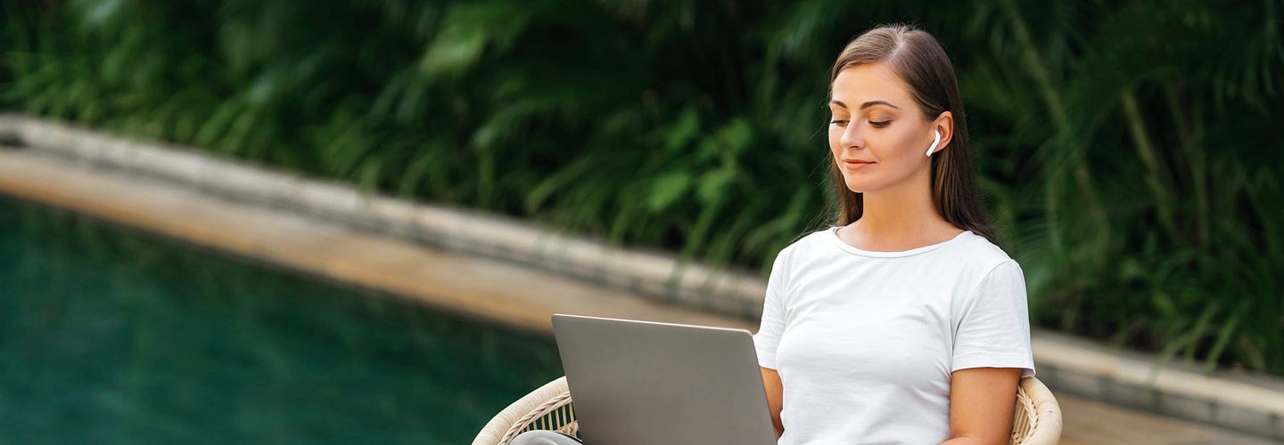 White woman in a white T-shirt and taupe pants sits cross-legged in a rattan chair with a laptop open in her lap and ear buds in her ears. Behind her is a green pool flanked by lush vegetation. She has straight brown hair.