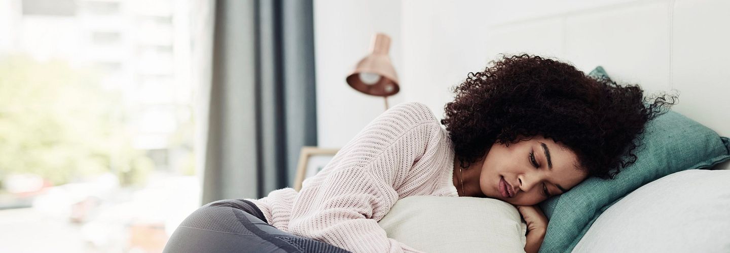 An African American woman in her 20s or 30s curls up in bed against a green throw pillow. She is wearing a light pink sweater and gray sweatpants. Her eyes are closed, and she is clutching a white pillow as if she is in discomfort.