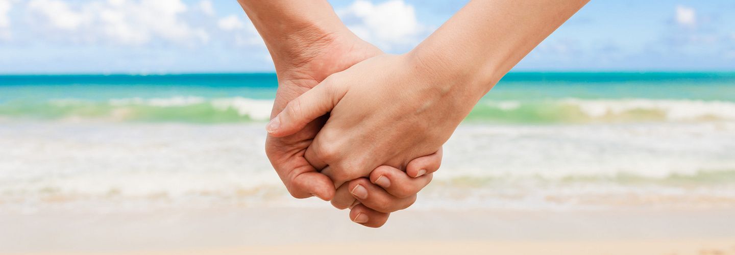 Closeup of a two hands holding each other with the ocean in the background on a sunny day.